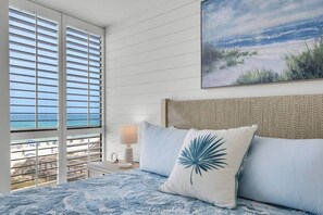 A bedroom with a view of the beach, a bed with blue bedding, a lamp, and a painting of a beach scene. - Oceanfront, Penthouse,Pool, Hot Tub, Elevator, Beach Service Included (Miramar Beach)