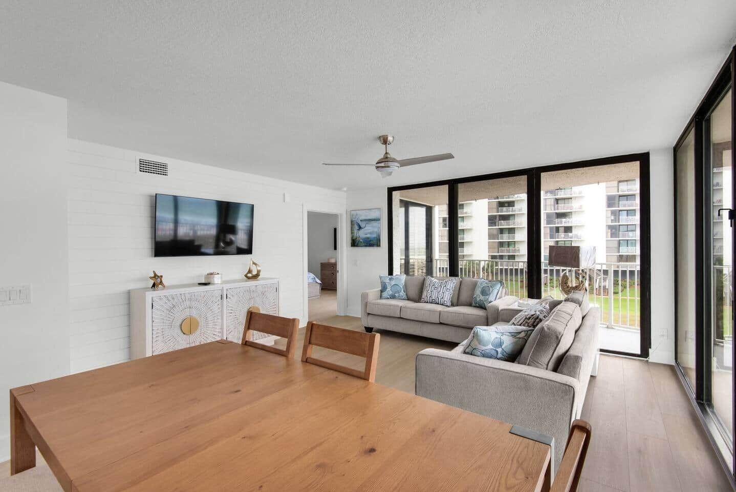 A modern living room with a wooden dining table, a sofa, and a TV mounted on the wall.
