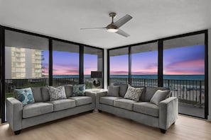 Ein Balkon mit Blick auf das Meer und einen Sonnenuntergang, zwei graue Sofas mit gemusterten Kissen, ein Deckenventilator und ein kleiner Tisch mit einer Lampe.