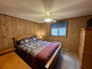 Ein Schlafzimmer mit einem hölzernen Kopfteil, einem Bett mit gemusterten Betttextilien, einem Deckenventilator, einem Fenster mit Vorhängen und einer Holzkommode.