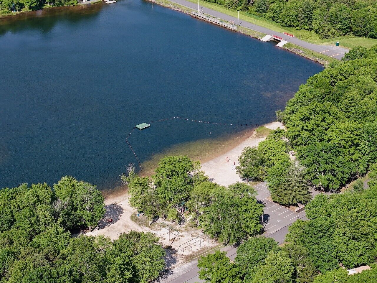Vista aerea di un lago circondato da vegetazione lussureggiante, una spiaggia sabbiosa e un'area parcheggio.