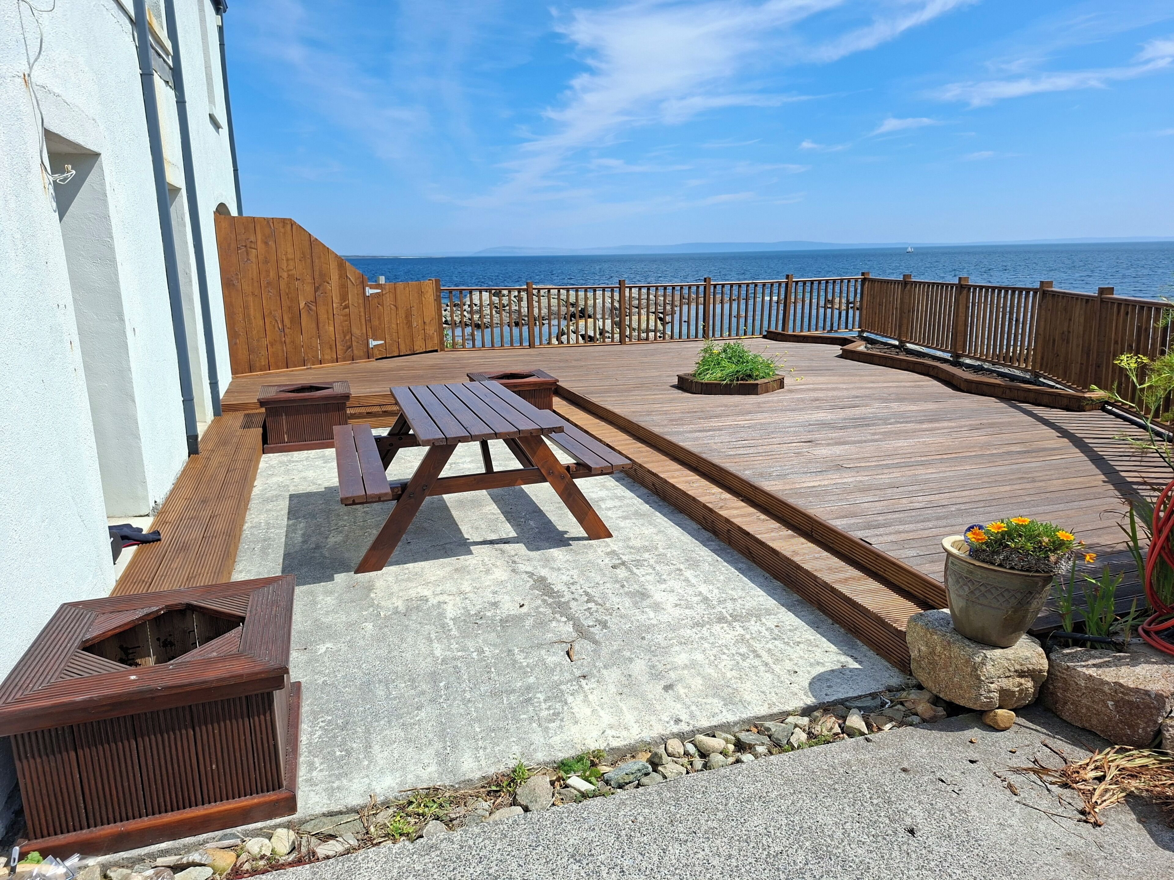 Eine Holzterrasse mit Picknicktischen und Pflanzkübeln, mit Blick auf ein Gewässer.
