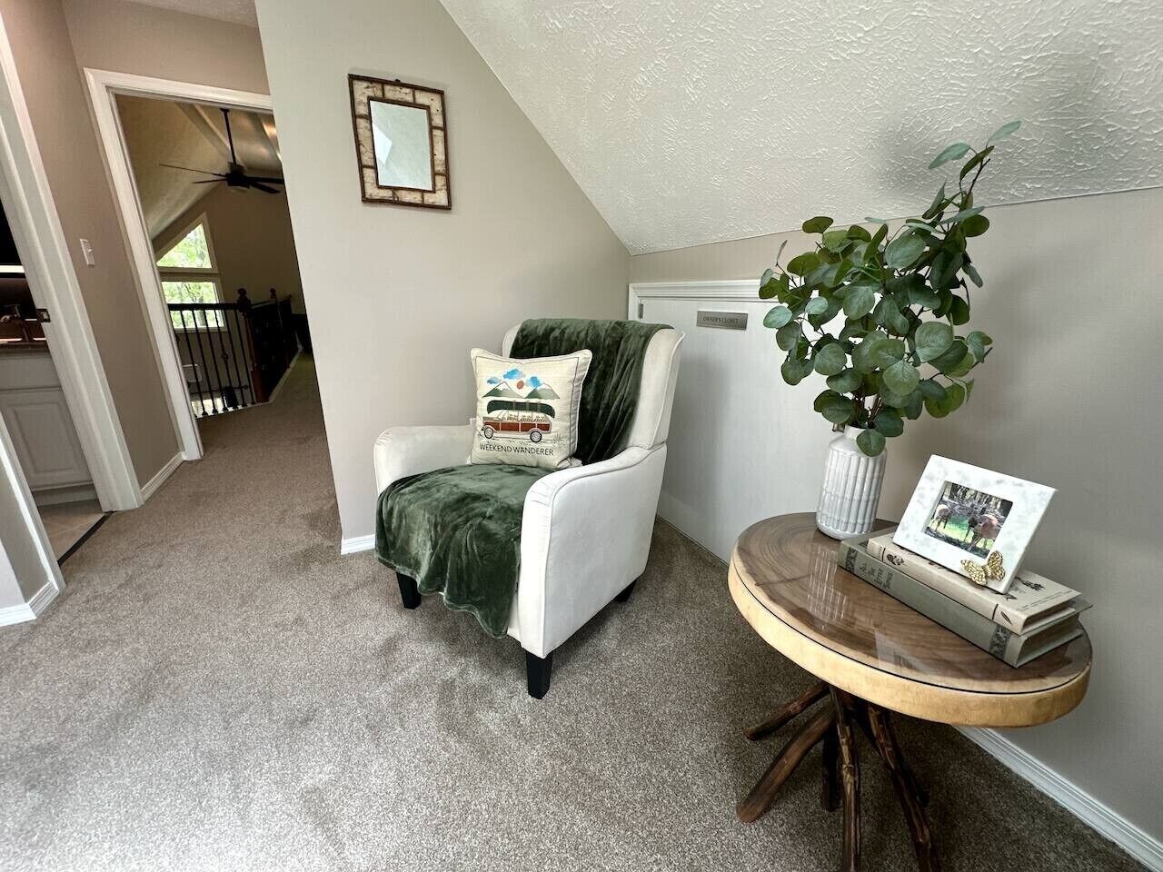 Ein Zimmer mit einem weißen Stuhl, einem kleinen runden Tisch mit Büchern und einem Bilderrahmen, einer grünen Pflanze und einem Deckenventilator.
