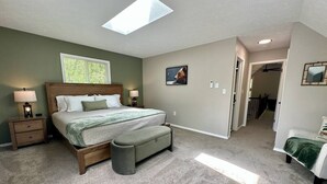Ein Schlafzimmer mit Bett, Nachttischen, einer Bank, einem Oberlicht und einem Flur, der in einen anderen Raum führt.