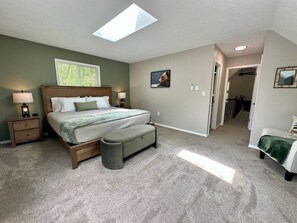 Ein Schlafzimmer mit Bett, Nachttischen, einer Bank, einem Oberlicht und einem Flur, der in einen anderen Raum führt.
