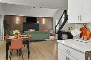 Una sala de estar moderna con chimenea, un comedor con mesa y sillas, y una cocina con armarios blancos y una tabla de cortar de madera.
