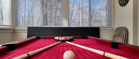 Une table de billard avec des billes et des queues, un jeu de fléchettes et une fenêtre avec des stores.