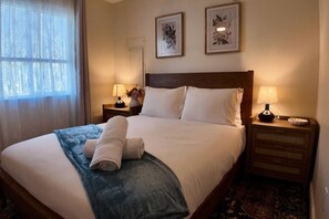 Ein ordentlich bezogenes Bett mit weißen Leinentüchern, zwei Nachttische mit Lampen und zwei gerahmte Blumenbilder an der Wand.