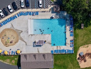 Vista aérea de una zona de piscina con sillones de descanso, una pequeña playa y un estacionamiento.