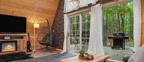 A cozy cabin interior with a fireplace, a hanging chair, a wooden coffee table, and a large window overlooking a forest.