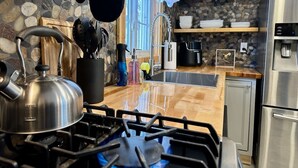 Une cuisine équipée d’une cuisinière à gaz, d’une bouilloire et de divers ustensiles. On y trouve un évier, un réfrigérateur et des étagères remplies de vaisselle.