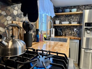 A kitchen with a gas stove, kettle, and various utensils. There is a sink, a refrigerator, and shelves with dishes. - Romantic A-Frame - Hot Tub - Pets Welcome (Blakeslee)