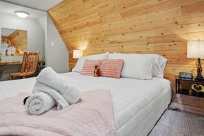A bedroom with a wooden sloped ceiling, a bed with white bedding, a bedside table with a clock, and a mirror on the wall. - Romantic A-Frame - Hot Tub - Pets Welcome (Blakeslee)