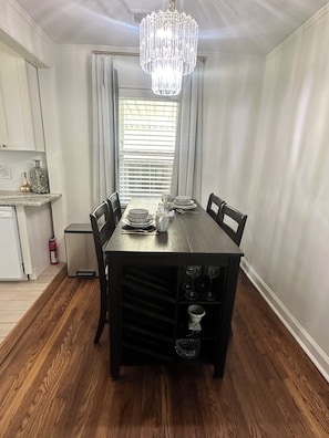 A dining area with a wooden table, chairs, a chandelier, and a window with blinds.