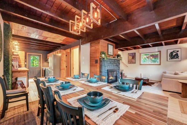Un espace repas avec une longue table en bois dressée pour six personnes, une cheminée et un coin salon confortable avec un canapé et un téléviseur.