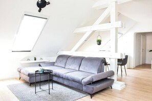 Une chambre moderne de style loft, avec un canapé d’angle gris, une petite table ronde et un plafond blanc aux poutres apparentes.