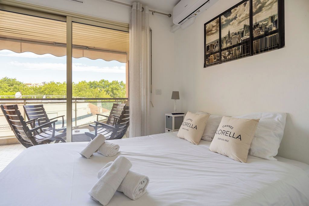 A hotel room with a bed, two pillows, a nightstand, a lamp, a cityscape painting, and a balcony with chairs.