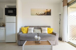 A living room with a grey sofa, two yellow pillows, a white vase with flowers, a painting on the wall, a kitchen area with a microwave and refrigerator, and a window with a view. - L'Aparté Cannois avec balcon en centre-ville (Cannes)