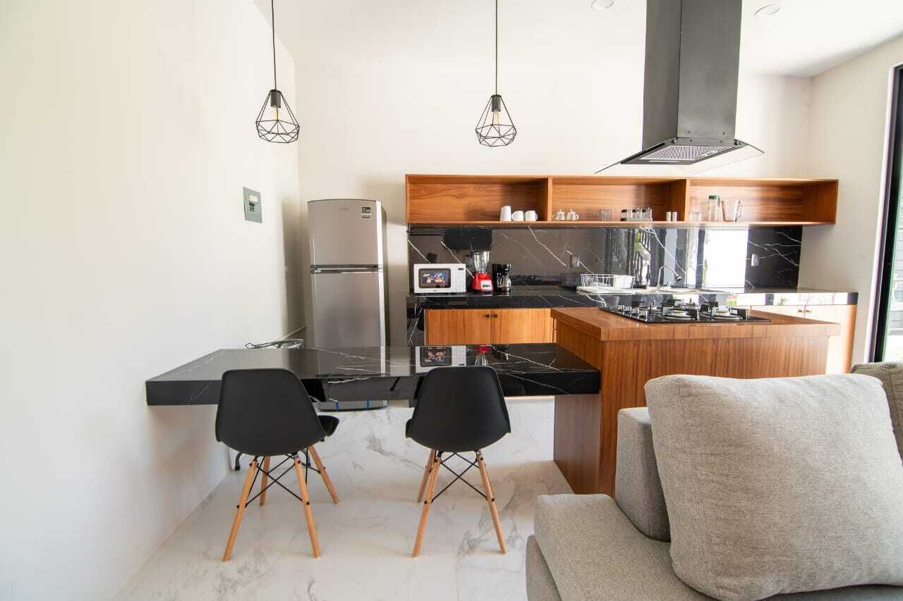 A compact kitchen with a refrigerator, microwave, and modern appliances. There are two black chairs and a sofa in the dining area.