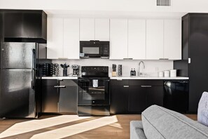 Una cocina moderna con electrodomésticos de acero inoxidable, armarios blancos y un sofá gris en la sala de estar.