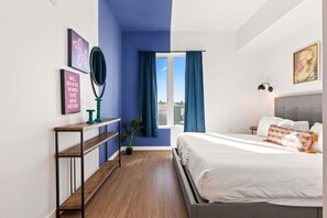 A bedroom with a bed, a nightstand, a mirror, a window with curtains, a wall with artwork, and a plant.