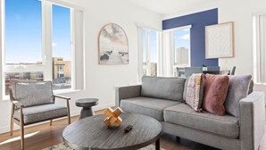 Ein modernes Wohnzimmer mit einem grauen Sofa, einem runden Couchtisch und Blick auf die Stadt durch große Fenster.