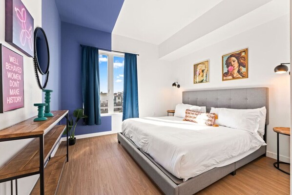 A modern bedroom with a large bed, a desk, a window with curtains, and artwork on the walls.