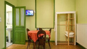 Une pièce avec une porte verte, un téléviseur fixé au mur, une table recouverte d’une nappe rouge, deux lits avec une literie bleue et une étagère en bois.