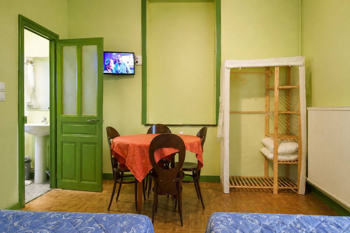 A room with a green door, a television mounted on the wall, a table with a red tablecloth, two beds with blue bedding, and a wooden shelf unit.