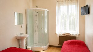 Une pièce avec une douche en verre, un lavabo, un lit recouvert d’une courtepointe rouge et une fenêtre avec un rideau.