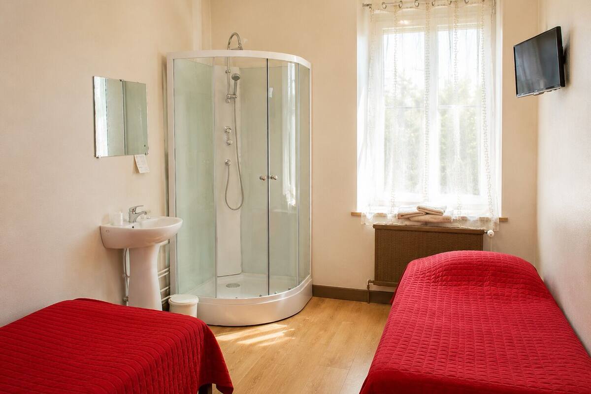 A room with a glass-enclosed shower, a sink, a bed with a red bedspread, and a window with a curtain.