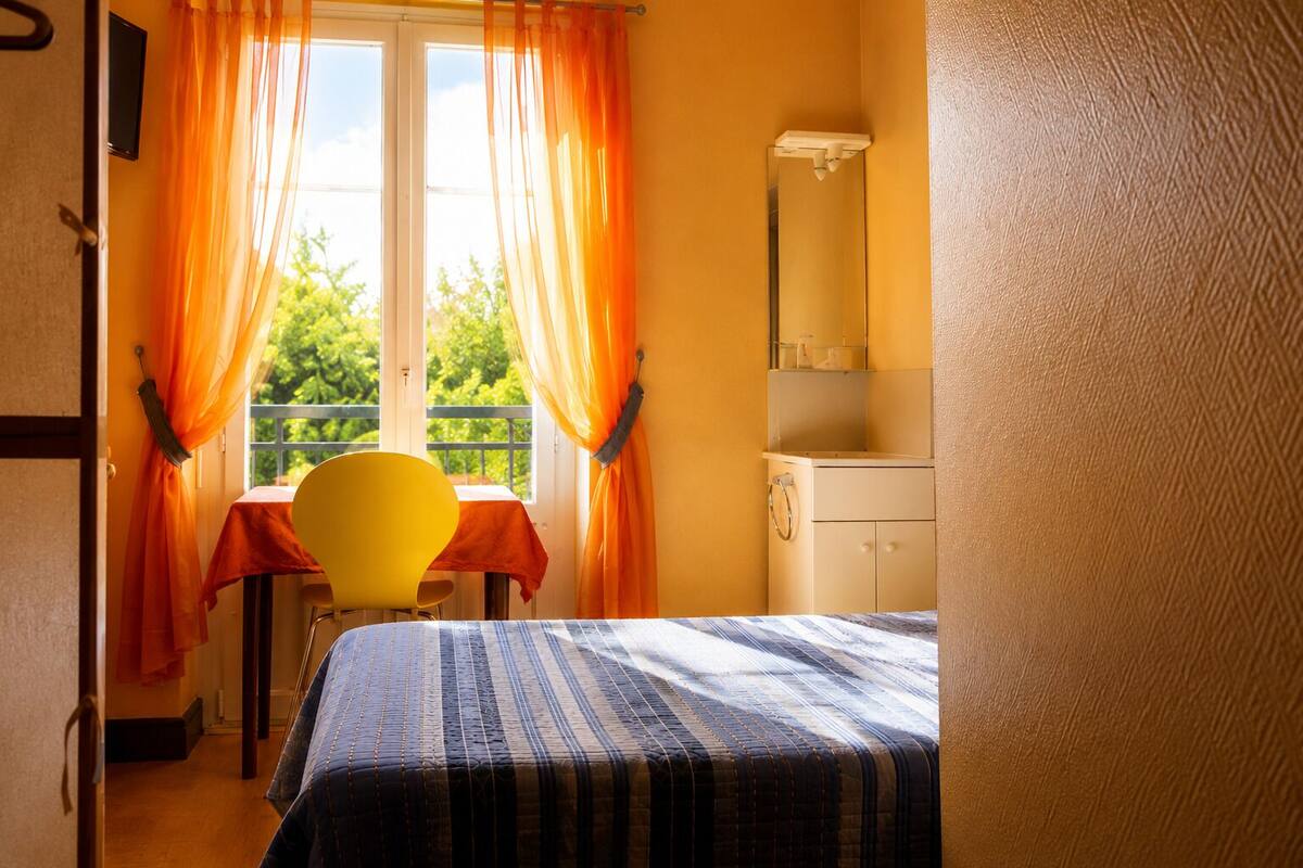 A room with a bed, a table, a chair, a window with curtains, a mirror, and a small cabinet.