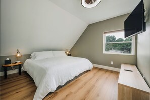 Ein Schlafzimmer mit einem Bett, Nachttischen, einer Lampe, einem an der Wand montierten Fernseher und einem Fenster mit Blick auf Grünflächen.