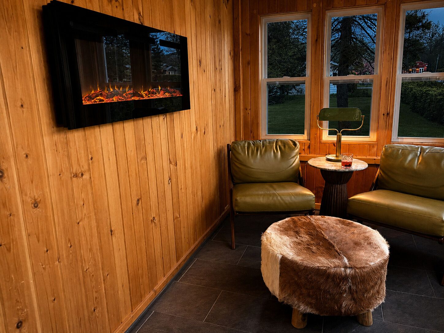 Una habitación acogedora con chimenea, dos sillones de cuero, una mesa redonda con lámpara y un otoman de piel.