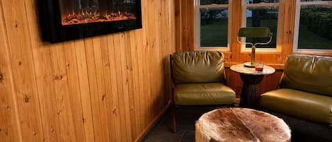 Una habitación acogedora con chimenea, dos sillones de cuero, una mesa redonda con lámpara y un otoman de piel.