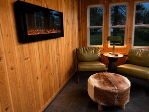 Ein gemütlicher Raum mit Kamin, zwei Ledersesseln, einem runden Tisch mit Lampe und einem Fellhocker.