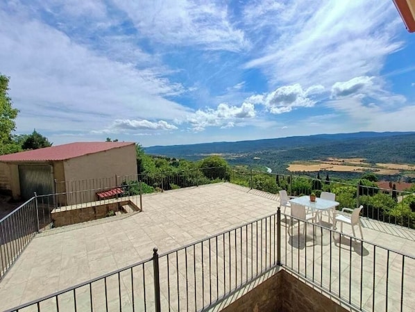 A terrace with a table and chairs, a small building with a red roof, and a scenic view of the landscape.