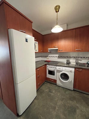 A kitchen with wooden cabinets, a white refrigerator, a stove, a washing machine, and a ceiling light fixture.