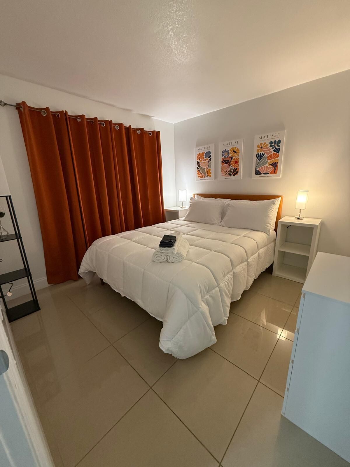 A neatly arranged bedroom with a large bed, a nightstand, a lamp, and a shelf with a folded towel.
