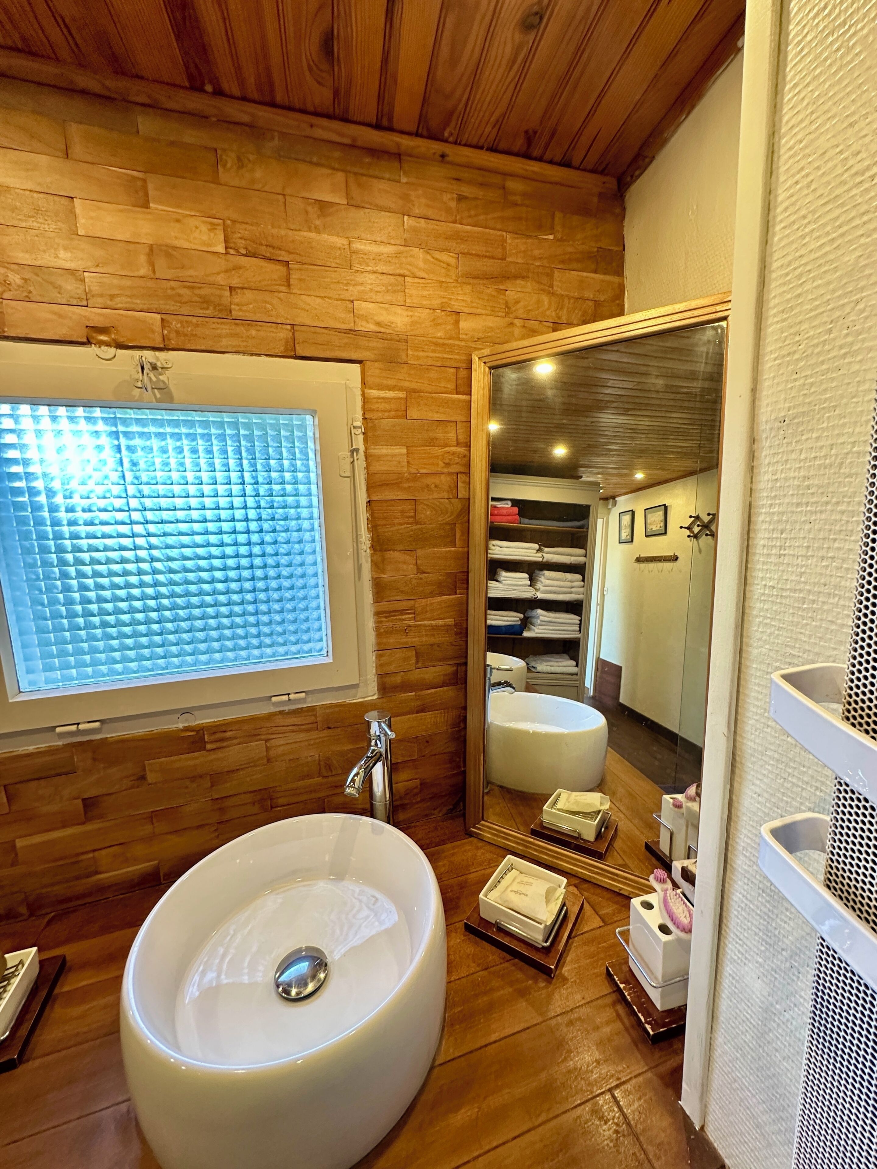 Un baño con un lavamanos ovalado grande, una ventana con persianas, un espejo y paredes de madera.