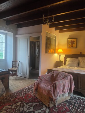 Ein Schlafzimmer mit Bett, Sessel, einer Holzkommode, einem gemusterten Teppich, einer Tür zu einem Badezimmer und einer Kronleuchter.