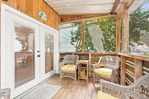 A covered porch with wicker furniture, a wooden table, and a glass door. - Coco Cottage w Outdoor Bar and TV Walk to Beach (North Myrtle Beach)