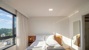 A modern hotel room with a large window, a bed with white linens, a bedside table with a lamp, and a view of the cityscape. - Studio 500m fr/ Vila Germanica, Blumenau Pin1907 (Blumenau)