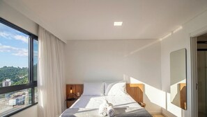 Quarto de hotel moderno com uma grande janela, cama com lençóis brancos, mesa de cabeceira com abajur e vista para o horizonte urbano.