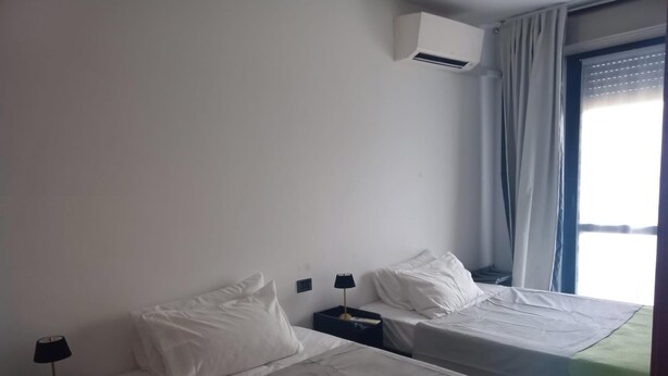 Quarto de hotel com duas camas de solteiro, mesa de cabeceira com abajur, ar condicionado e janela com cortinas.