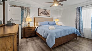 Une chambre à coucher avec un lit en bois, deux tables de chevet avec des lampes, un ventilateur de plafond, un tableau encadré au mur et une fenêtre avec des rideaux.