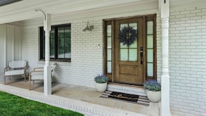 Une maison en briques blanches avec une porte d’entrée en bois, deux plantes en pot, un banc et un paillasson.