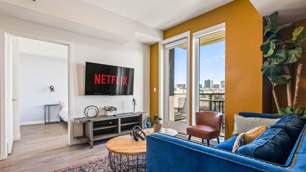 A living room with a large TV, a sofa, a coffee table, and a view of the city through large windows.