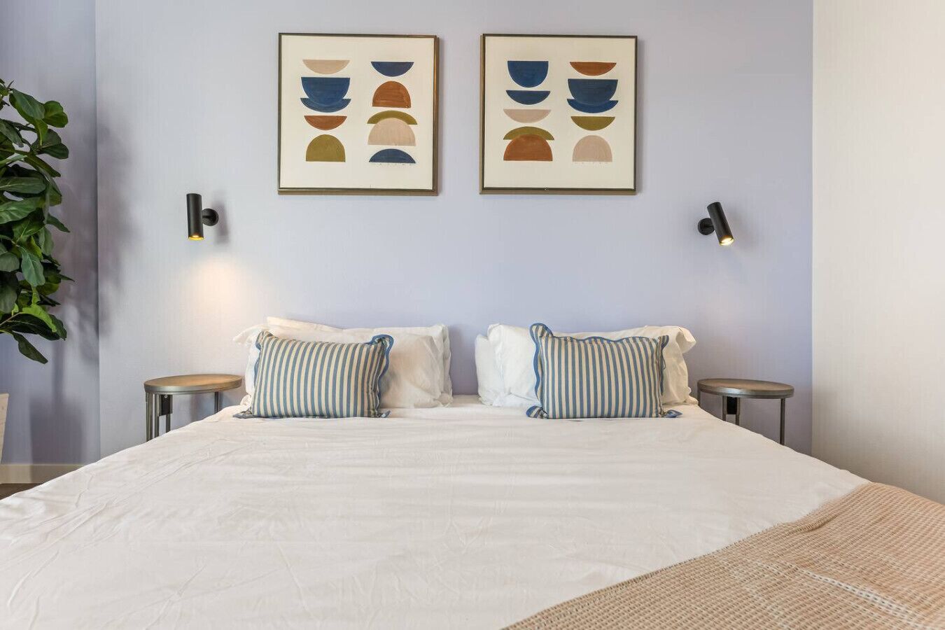 Una cama doble con dos mesitas de noche, un aplique de pared y dos cuadros abstractos enmarcados sobre la cama.