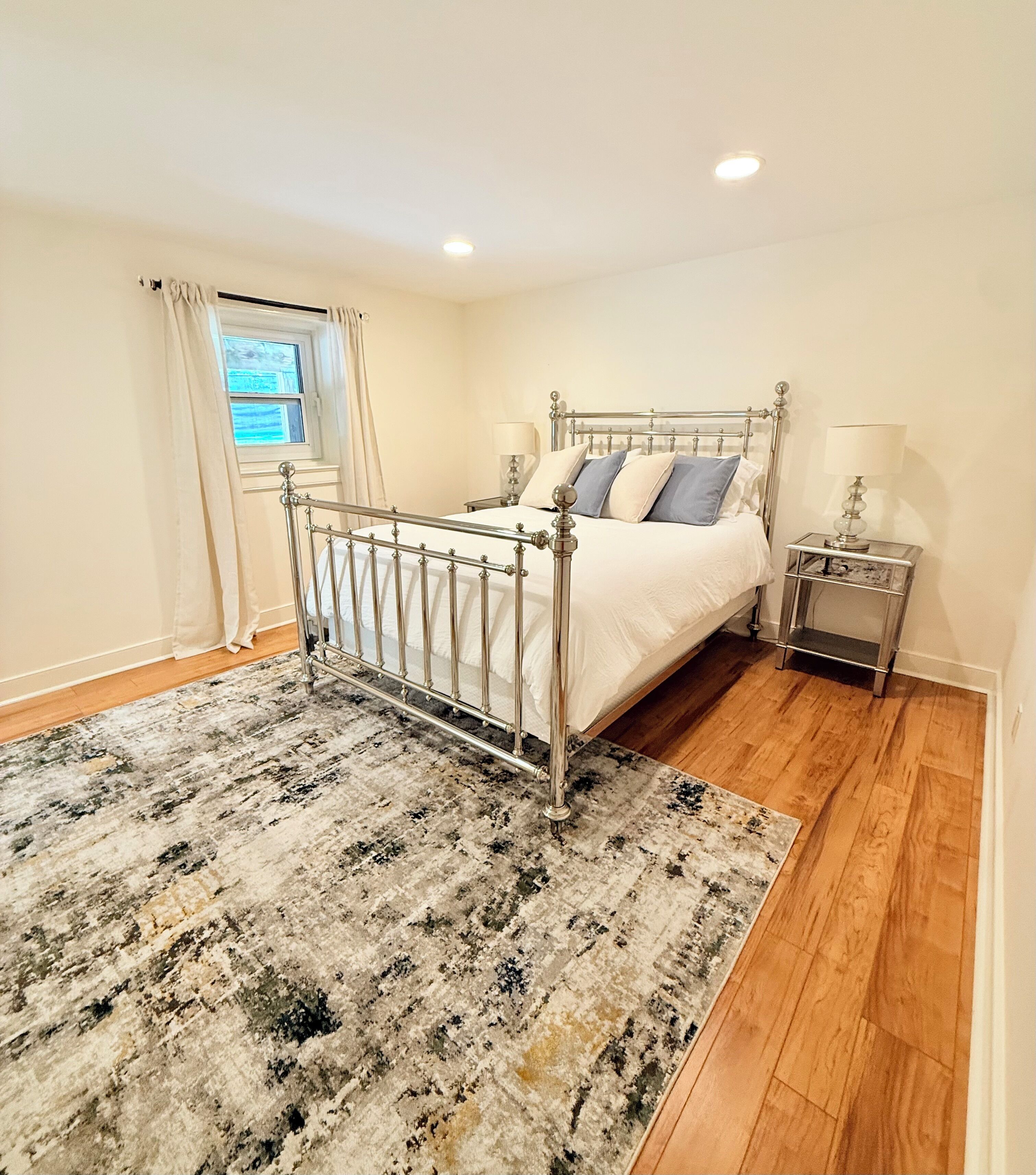 A bedroom with a metal-framed bed, a bedside table with a lamp, a window with curtains, and a patterned rug.